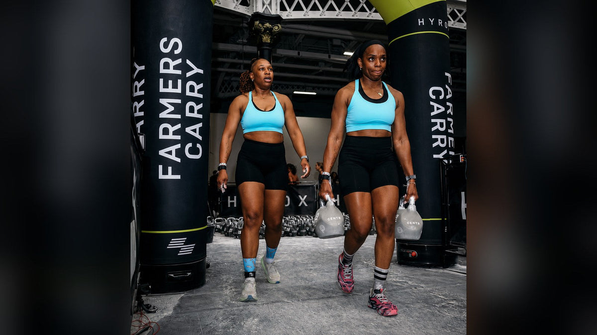 Due atlete in gara HYROX durante il farmers carry con i kettlebell, con calzini HEXXEE ben visibili