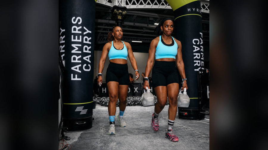 Due atlete in gara HYROX durante il farmers carry con i kettlebell, con calzini HEXXEE ben visibili